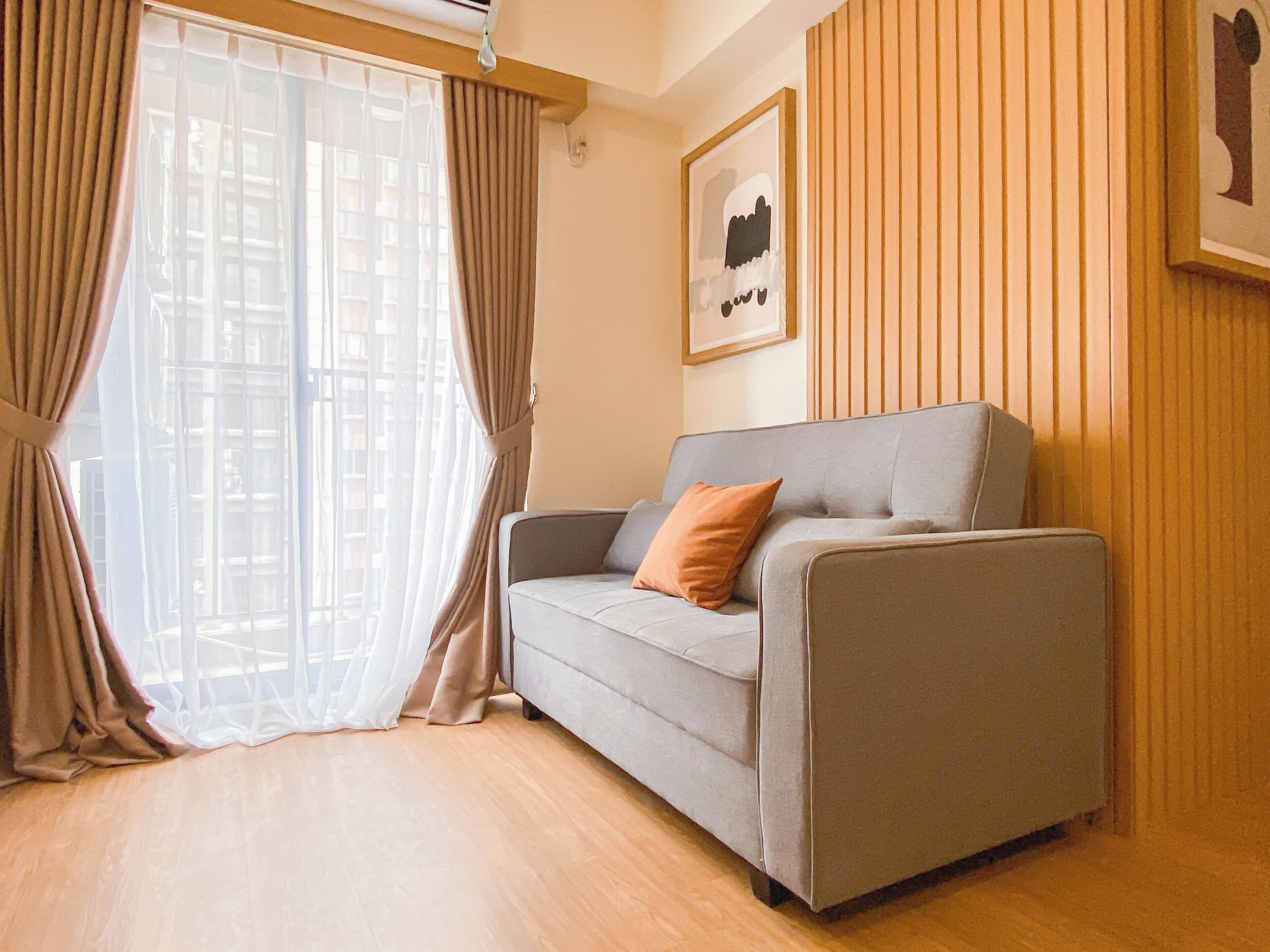 A modern living room with a grey sofa, a single orange pillow, and a large window with sheer curtains.