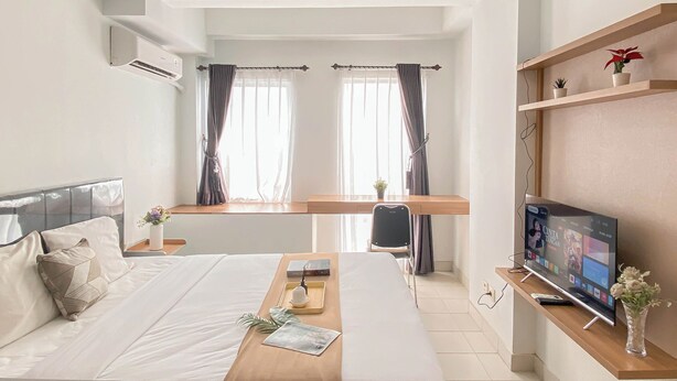 A modern hotel room with a large bed, a desk with a chair, a TV, and a small shelf with plants. - Standard Look Studio At Patraland Urbano Apartment (Bekasi)