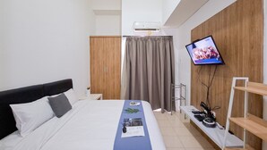 A hotel room with a bed, a TV mounted on the wall, a wooden wardrobe, and a desk with a computer.