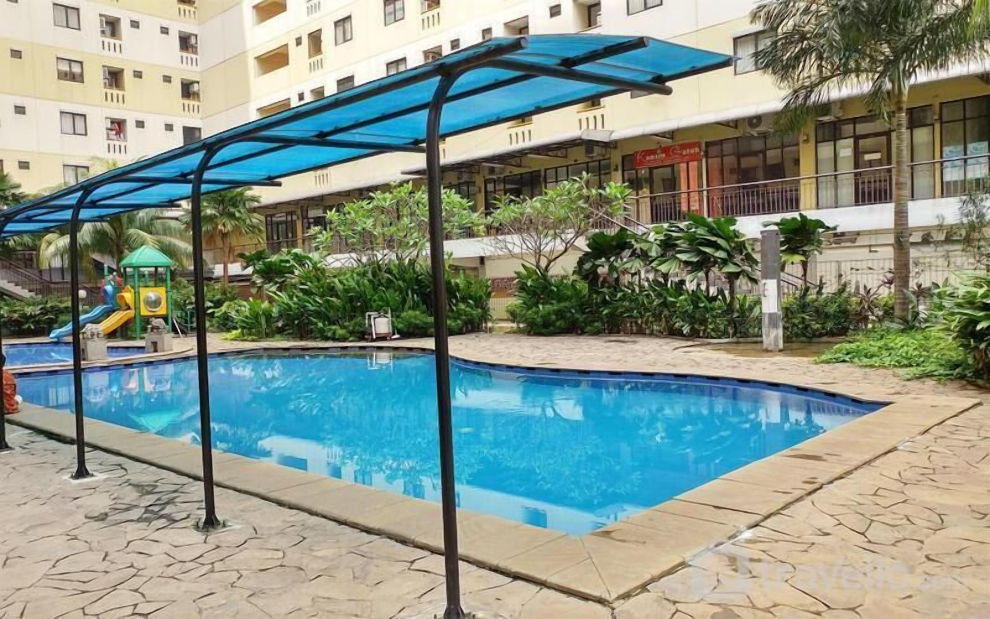 Une piscine surmontée d’un auvent bleu, un espace de jeux pour enfants et, au fond, un bâtiment doté de balcons.