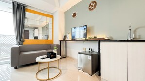 A compact living space with a sofa, a small round table, a refrigerator, and a TV mounted on the wall. - Modern And Tidy Studio Room At West Vista Apartment (Jakarta)