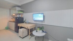 A small kitchenette with a refrigerator, microwave, and a coffee maker. There is a TV mounted on the wall, a small table with a plant, and a white chair. - Spacious And Modern Studio At Tanglin Supermall Mansion Apartment (Surabaya)