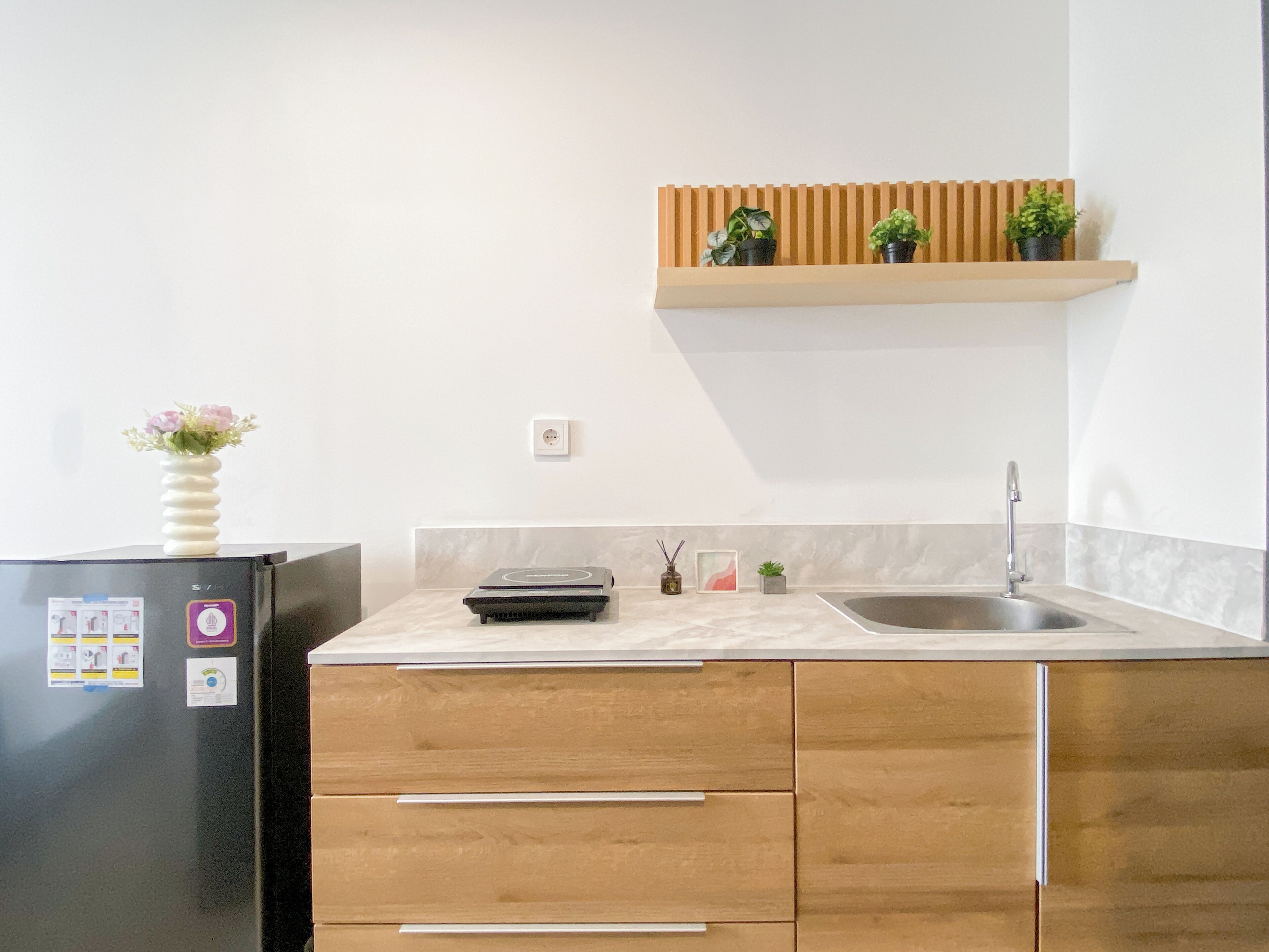 A kitchen with a black refrigerator, wooden cabinets, a marble countertop, a wooden shelf with potted plants, a sink, and a vase with flowers on the fridge.