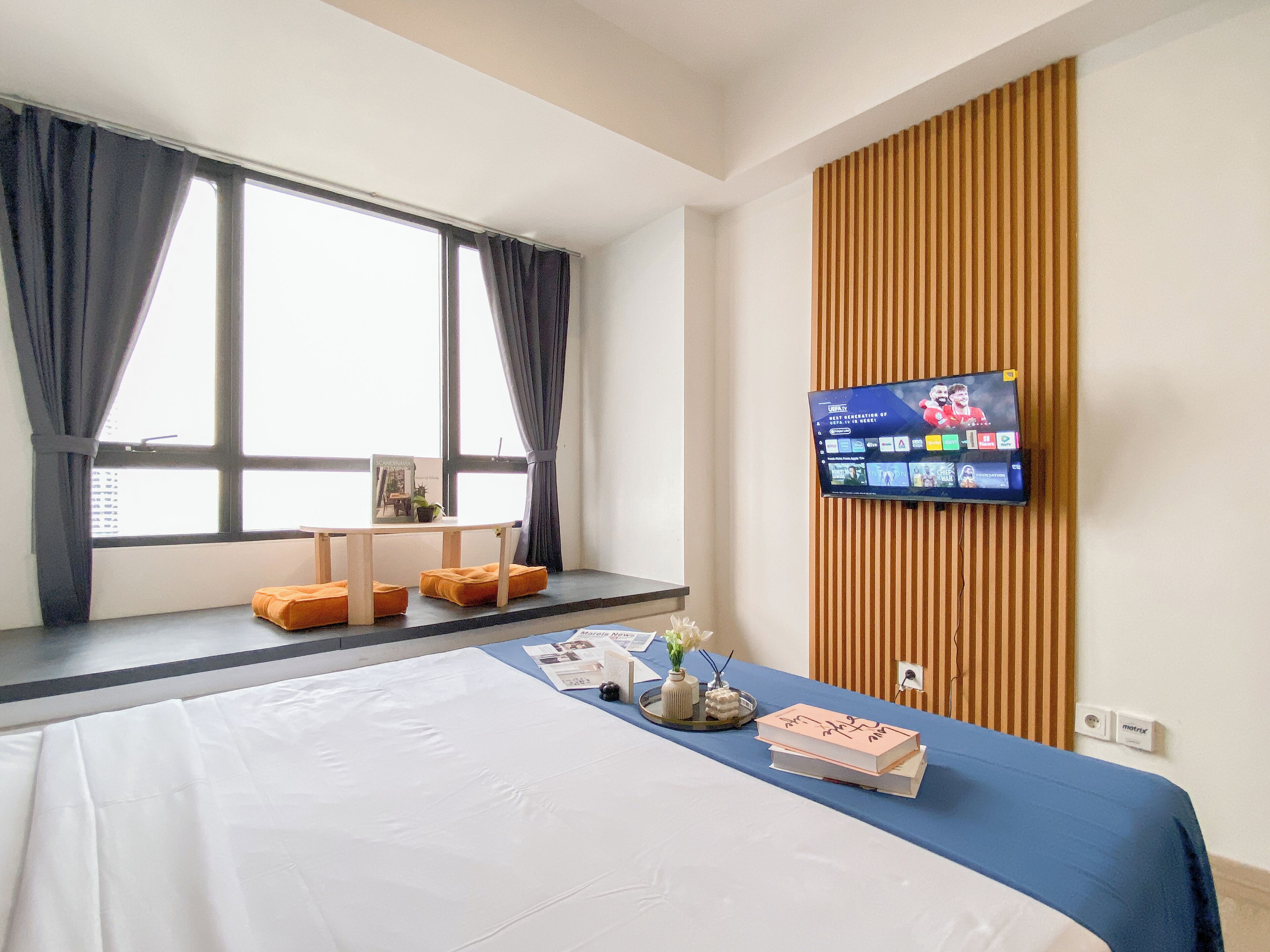 Hotelkamer met een groot bed, een televisie bevestigd aan een houten wandpaneel, een raam met gordijnen, en een klein tafeltje met een boek en decoratieve objecten.