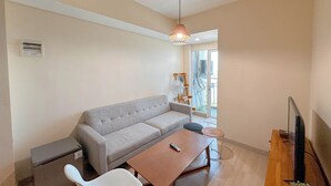 A modern living room with a grey sofa, a wooden coffee table, a TV on a wooden stand, and a ceiling fan. - 1Br Near Uinsa At Tamansari Papilio Apartment (Surabaya)