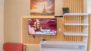 A modern living room with a flat-screen TV mounted on a wooden wall, a red polka-dotted towel, and a shelf with a book. - Pure Nest Studio At Candiland Apartment (Semarang)