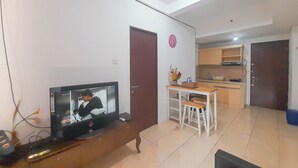 A living room with a television displaying a person, a dining area with a table and chairs, and a kitchen area with cabinets and a countertop. - 1Br Near Gedebage Station At Tamansari Panoramic Apartment (Bandung)
