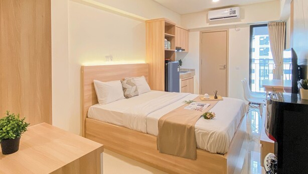 A modern hotel room with a wooden bed, a small kitchenette, a window with a view, and an air conditioning unit. - Warm Minimalist Studio Apartment Near Paramadina University At Meikarta (Cikarang)