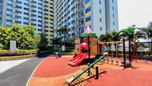 A playground with a slide, swings, and a climbing structure in front of a tall residential building. - Basic Studio Apartment At Springlake Summarecon Bekasi (Bekasi)