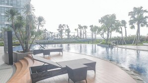 A poolside area with lounge chairs and a modern building in the background. - Modern And Stunning 2Br At Anderson Supermall Mansion Apartment (Surabaya)