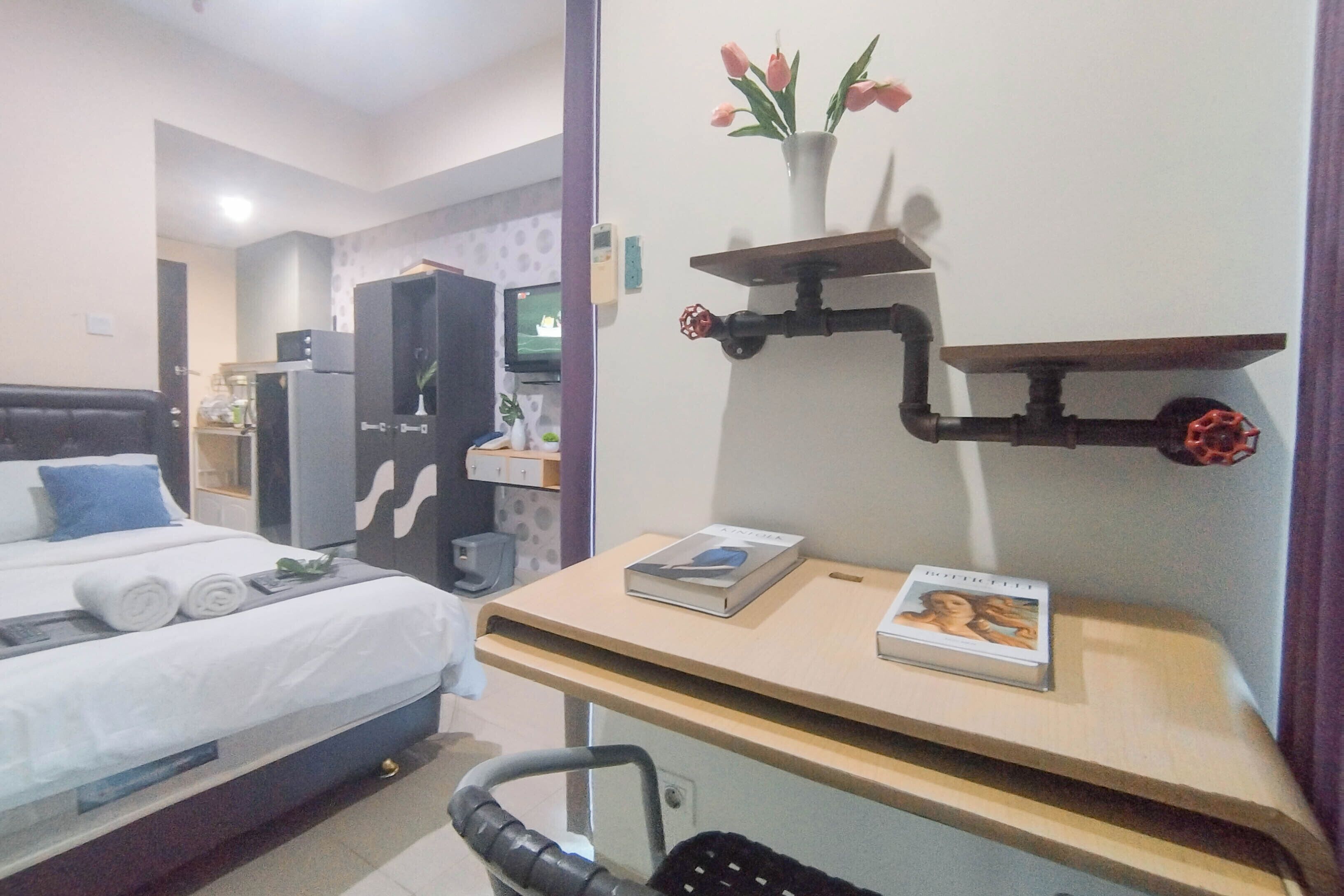 Ein Hotelzimmer mit Bett, Schreibtisch mit Büchern, einer Wandablage mit Blumen und einer Dusche.