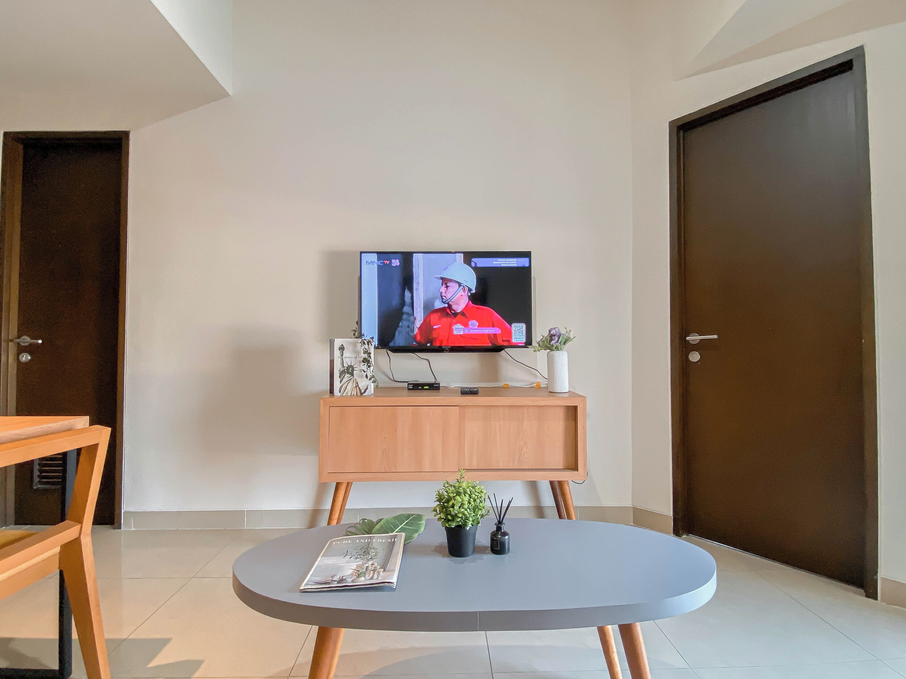 Un salon moderne avec un téléviseur posé sur un meuble en bois, une table basse ronde sur laquelle se trouvent un magazine et une plante, et une porte sur le côté droit.
