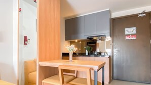 A compact kitchen with a dining area, including a wooden table and chairs, a vase with flowers, and a brown door with a fire safety sign. - 1Br Apartment Near President University At Mustika Golf Residence (Cikarang)