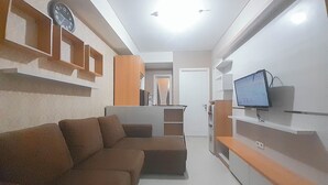 A modern living room with a brown sectional sofa, built-in shelving, a flat-screen TV, and a clock on the wall. - 2Br Near Unpar At Parahyangan Residence Apartment (Bandung)