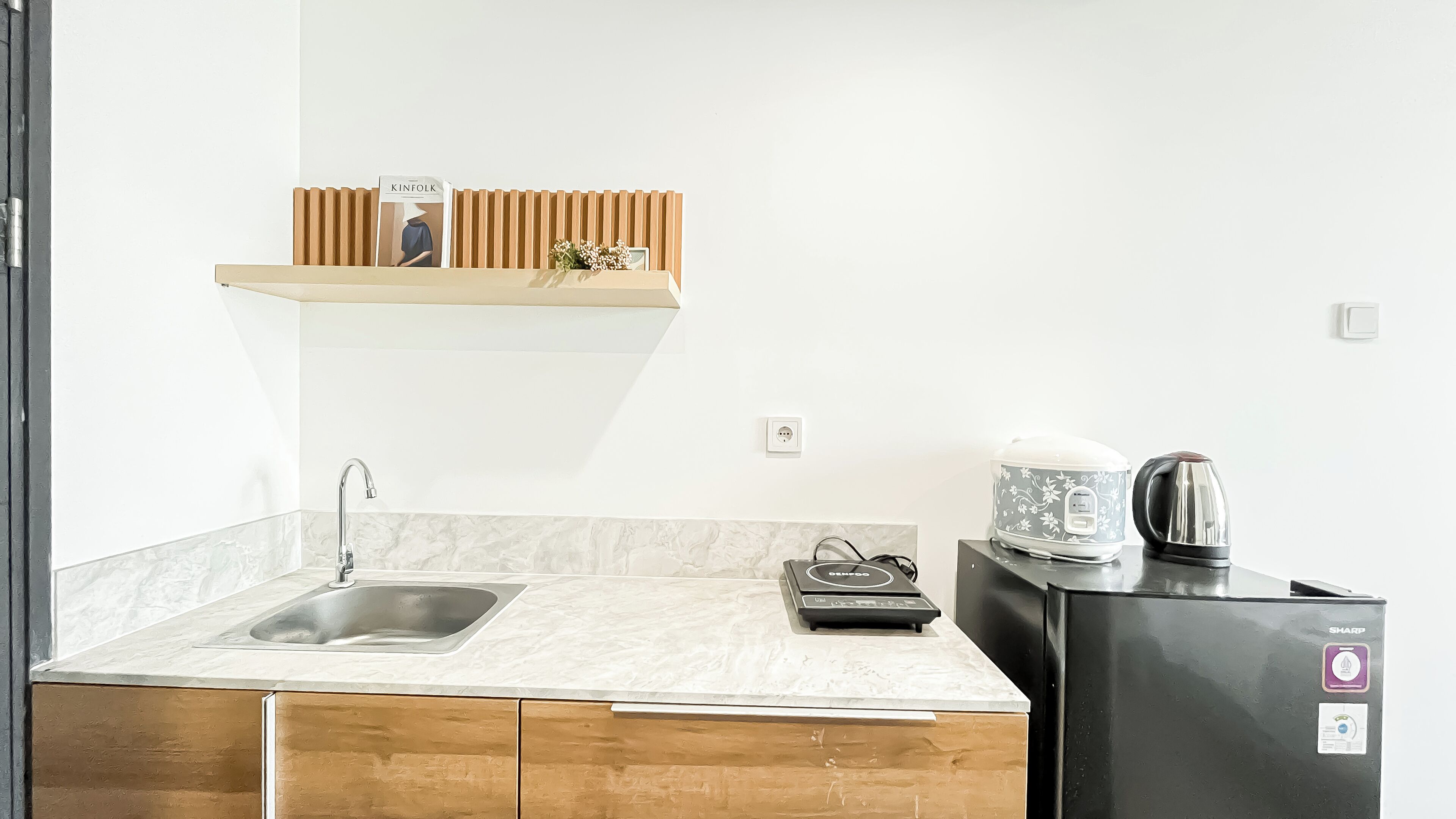 A kitchen with a sink, countertop, wooden cabinets, a kettle, a coffee maker, and a small shelf with a book.