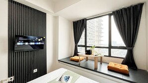 A modern hotel room with a flat-screen TV mounted on a wall, a small wooden table with a plant, and a window with black curtains. - Cozy Nordic Style Apartment At Collins Boulevard (Tangerang)