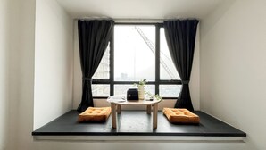 A small window seat with two cushions, a table with a vase and a remote control, and a view of a cityscape with a crane. - Cozy Nordic Style Apartment At Collins Boulevard (Tangerang)