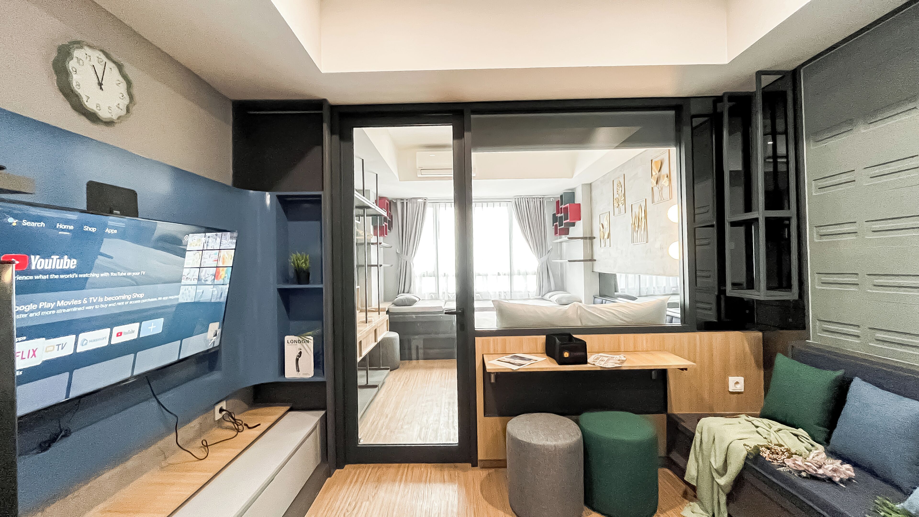 A modern hotel room with a large TV screen displaying YouTube, a wooden desk, a comfortable seating area with a sofa and ottoman, and a view into the adjacent room.