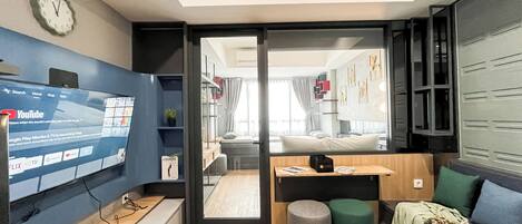 A modern hotel room with a large TV screen displaying YouTube, a wooden desk, a comfortable seating area with a sofa and ottoman, and a view into the adjacent room.