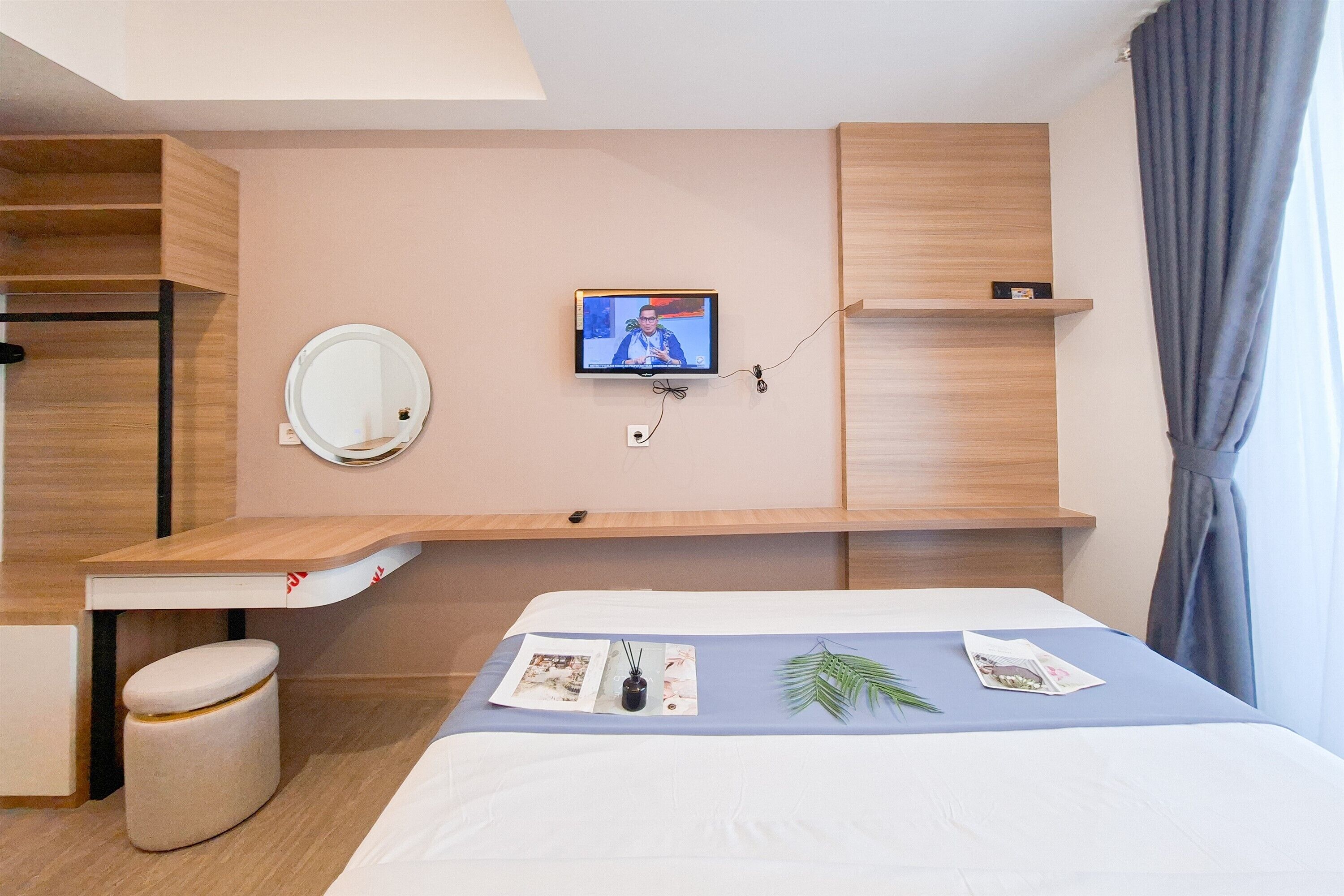 Ett hotellrum med en säng, en tv som är monterad på väggen, ett litet skrivbord, en spegel och ett fönster med gardiner.