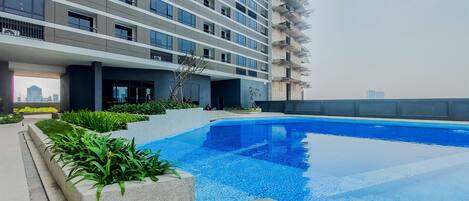 A modern building with a swimming pool, surrounded by greenery and a clear sky.