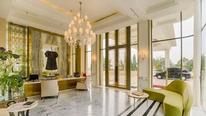 A modern hotel lobby with a marble floor, a chandelier, a reception desk, a green sofa, and large windows offering a view of the outside. - Minimalist Look Studio Apartment At Art Deco (Bandung)