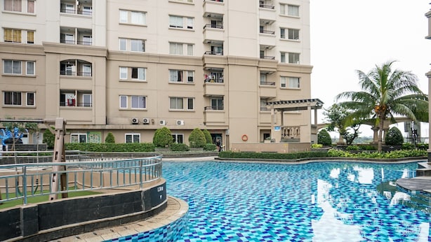 Una piscina con baldosas azules a cuadros, rodeada por un muro de ladrillo bajo. Al fondo, un edificio de varios pisos con balcones y ventanas.