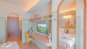 A compact hotel room with a bed, a desk with a chair, a TV mounted on the wall, and a small balcony area. - Chic Style Studio Apartment Grand Kamala Lagoon Bekasi (Bekasi)