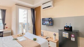 A hotel room with a bed, desk, and a TV mounted on the wall. There is an air conditioning unit above the bed and a refrigerator with stickers on it. - Basic Style Studio At Apartment Belmont Residence Puri (Jakarta)