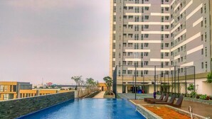Una piscina en la azotea con vistas a un rascacielos, rodeada de un área ajardinada con plantas y una zona de estar con sillones reclinables.