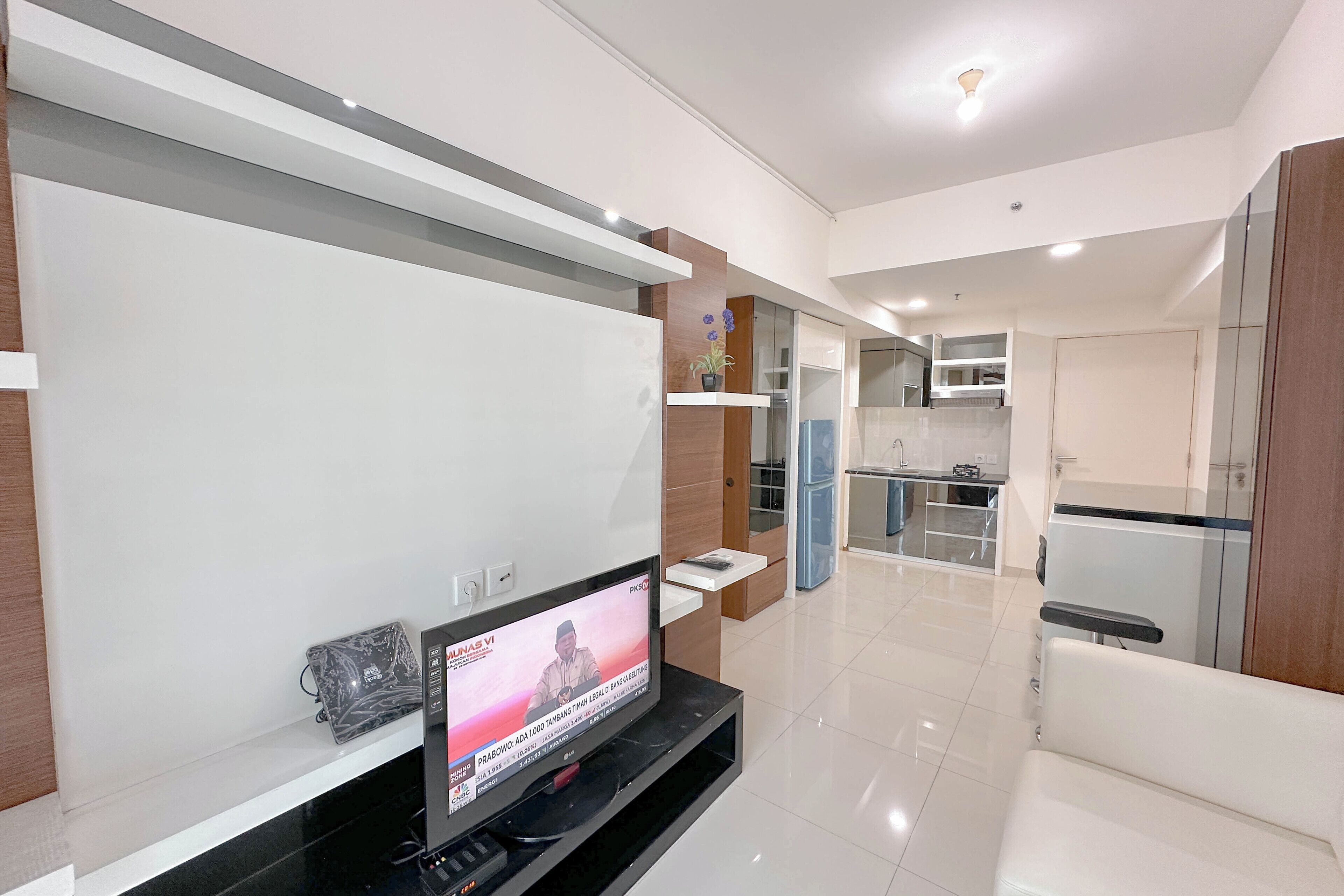 A modern living room with a flat-screen TV on a stand, a sofa, and a kitchen area with cabinets and a refrigerator.