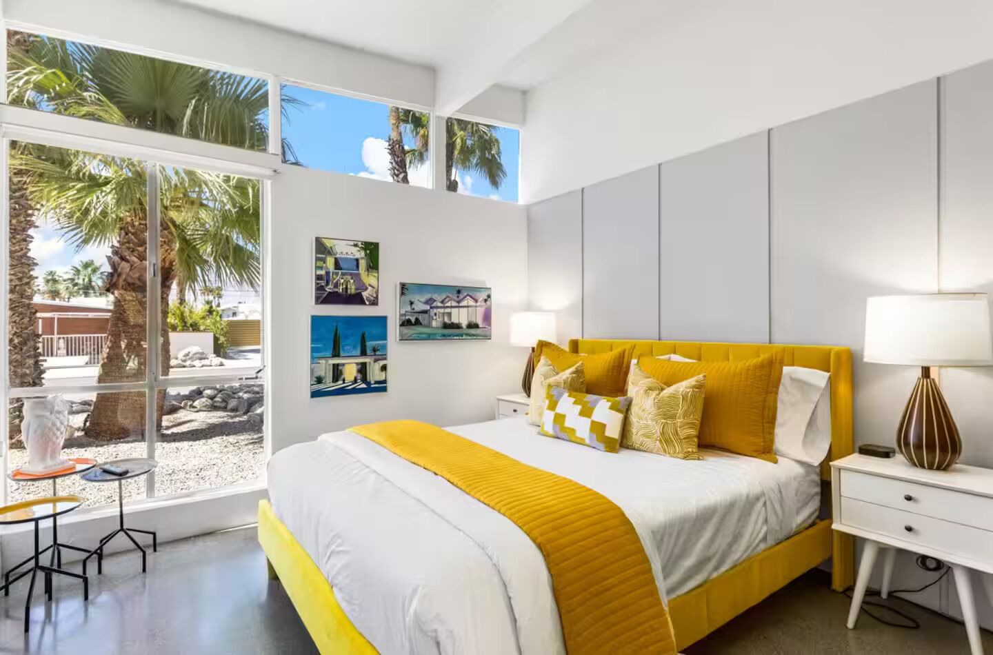 A modern bedroom with a large bed, a bedside table, a lamp, and a window with a view of palm trees.