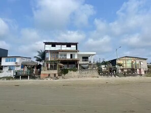 A two-story building with a flat roof and a balcony, surrounded by a sandy area and other buildings.
