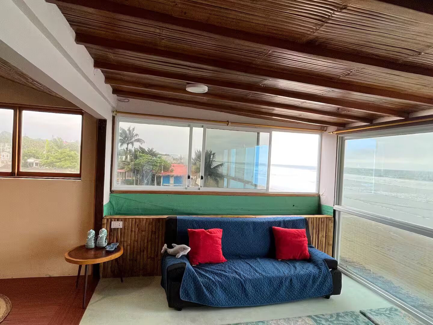 Une pièce avec un plafond en bois, un canapé bleu agrémenté de coussins rouges, une petite table en bois sur laquelle sont posés des salières et des poivrières, et une grande fenêtre donnant sur une plage.