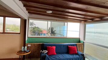Une pièce avec un plafond en bois, un canapé bleu agrémenté de coussins rouges, une petite table en bois sur laquelle sont posés des salières et des poivrières, et une grande fenêtre donnant sur une plage.