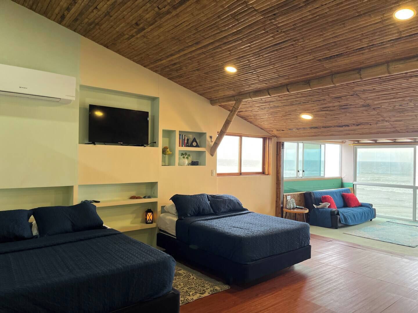 Habitación con dos camas, un televisor, estanterías con libros y un reloj, y vistas al mar a través de amplias ventanas.