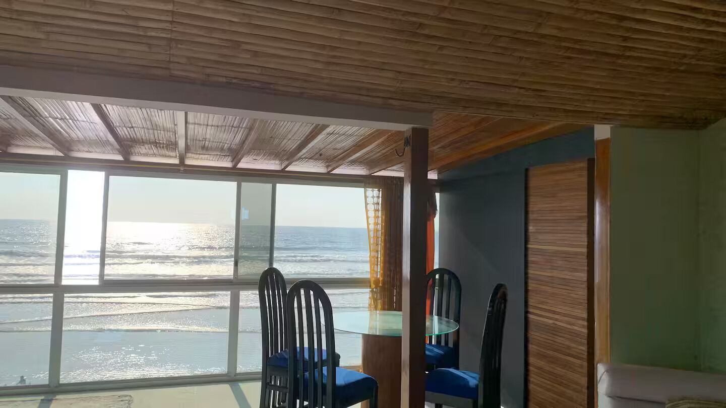 Une pièce dotée d’une grande fenêtre donnant sur l’océan, d’un plafond en bois et d’un coin repas avec des chaises et une table.