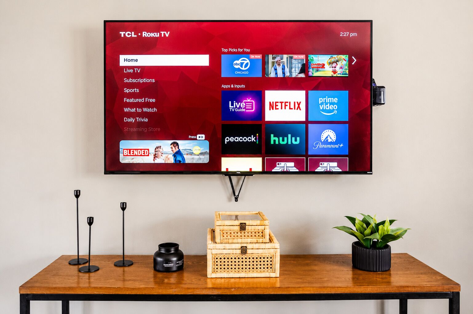A TCL Roku TV mounted on a wall displaying various streaming services and a table with a plant and a basket.