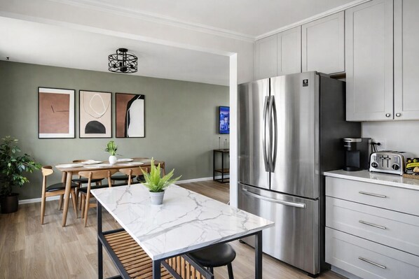 A modern kitchen with a stainless steel refrigerator, a dining area with a marble table, and a potted plant.