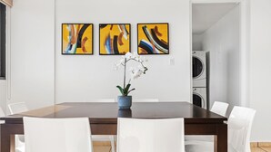 A dining area with a dark wooden table, white chairs, and three abstract paintings on the wall. - Waterside Flat With Garage and Easy Beach Access (San Juan)