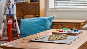 Une table en bois sur laquelle sont disposées des lettres de Scrabble formant les mots « BEACH BLISS » et d’autres mots, une échelle rouge et un coussin bleu, dans une pièce avec une fenêtre et une bibliothèque en arrière-plan.