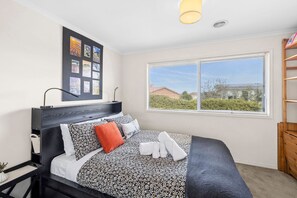 A bedroom with a bed, a bedside table, a lamp, a wall with pictures, a window with a view, and a wooden shelf.