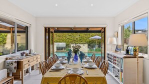 Un coin repas avec une table dressée pour quatre personnes, une piscine visible à travers la porte vitrée et un espace cuisine avec un meuble à vin.