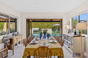 A dining area with a table set for four, a pool visible through the glass door, and a kitchen area with a wine rack. - Beach Living Bliss (Torquay)