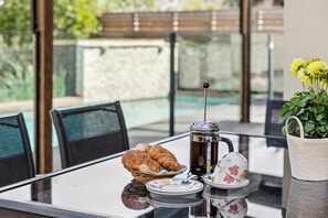 A table with croissants, a coffee pot, and a teapot, set against a glass table with a view of a pool and greenery. - Beach Living Bliss (Torquay)