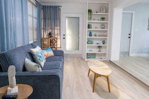 A living room with a grey sofa, a small wooden table, a wooden chair, a bookshelf, and a door.