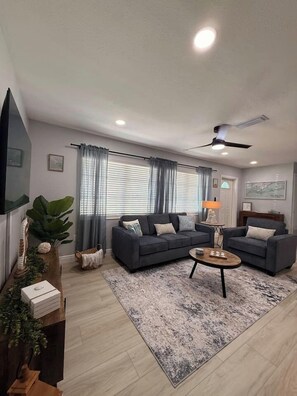A modern living room with a sofa set, a coffee table, a ceiling fan, and a large window with curtains.