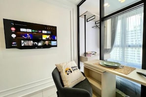 A hotel room with a flat-screen TV mounted on the wall displaying various channels, a desk with a plate and a cup, a chair with a pillow, and a window with a view of buildings.