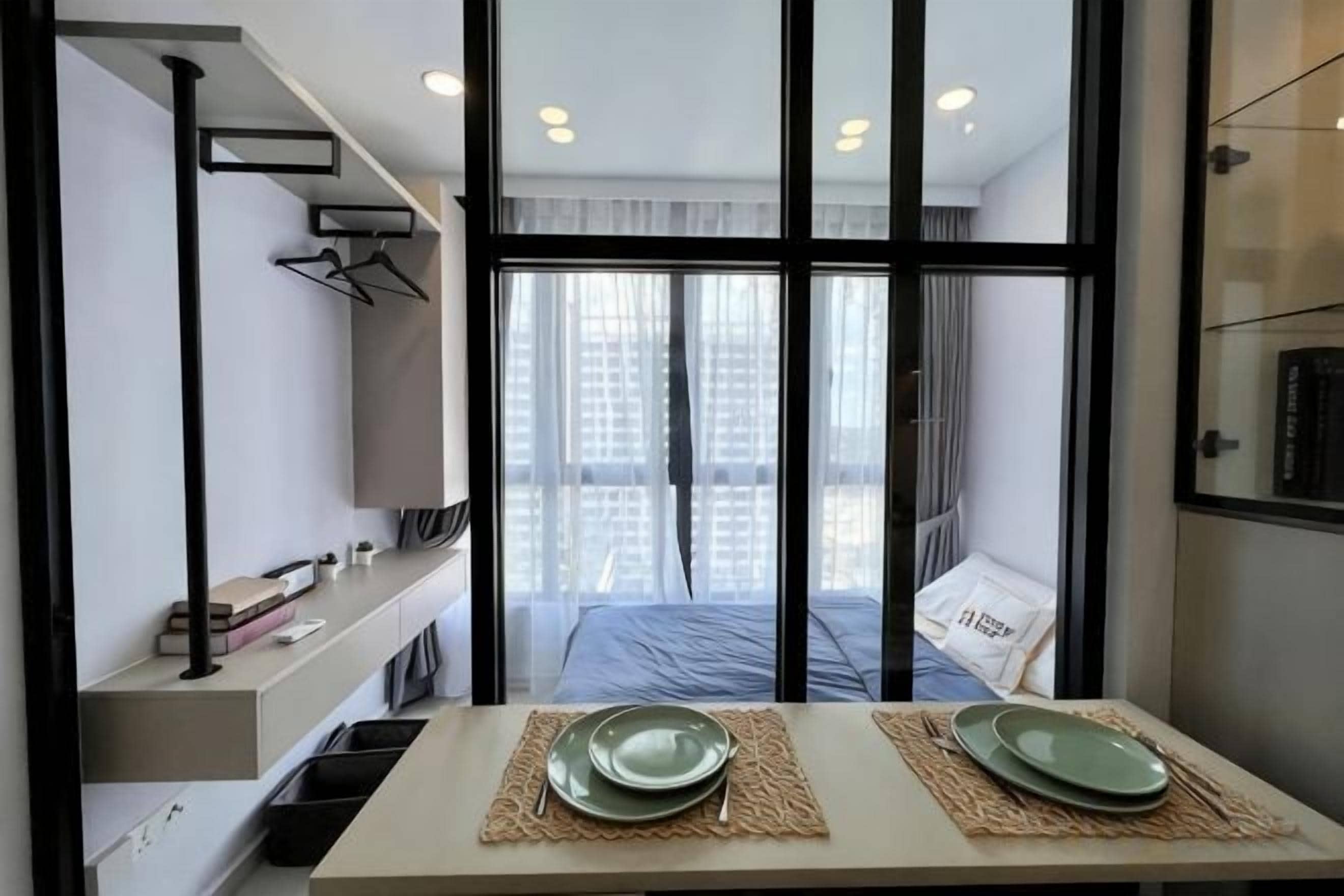 Una habitación de hotel moderna con un ventanal grande, una cama, una mesa con platos y una estantería con libros y una lámpara.