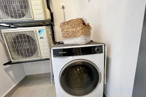 A front-loading washing machine with a basket on top, a wall-mounted air conditioner, and a laundry rack. - Gaia Residence, Gamuda Gardens Rawang by Sleepy Bear (Rawang)